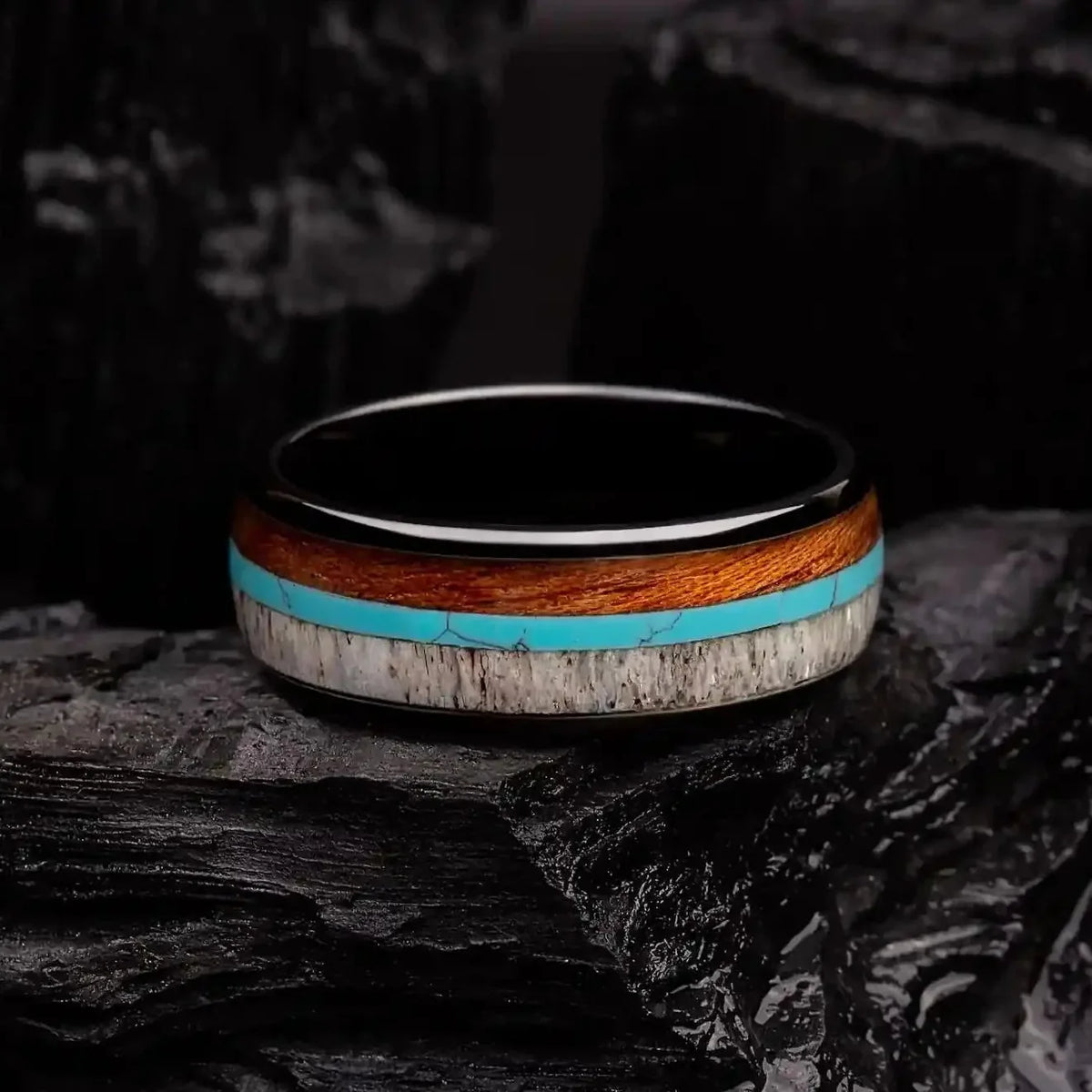 Brushed black tungsten ring with turquoise, koa wood and deer antler. The ring is laying on a black coal
